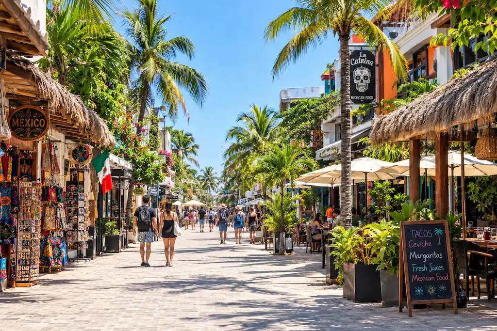 Bustling Quinta Avenida in Playa del Carmen, Mexico, lined with palm trees, colorful shops, Mexican souvenirs, and outdoor restaurants beneath a bright blue sky