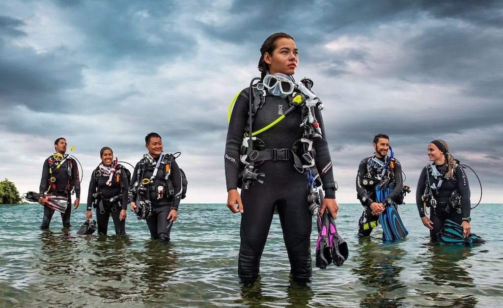 A group of Divemaster students standing in shallow ocean water, fully equipped with dive gear and fins, with one Divemaster standing prominently in the foreground under a cloudy sky.
