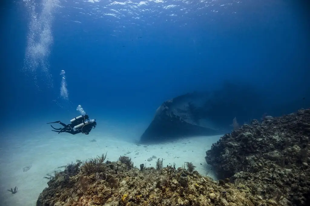 Advanced scuba diver exploring a clear blue underwater reef, swimming above a sandy seabed beside coral formations and a large submerged rock.