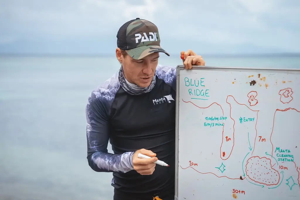 Scuba diving professional giving a dive briefing beside a whiteboard showing a hand-drawn dive site map, including depth markings, a safety stop, entry point, with the ocean in the background.