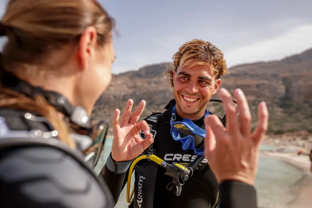 Scuba instructor and student in wetsuits giving the “OK” hand signal after a successful training dive, with a rocky coastline in the background