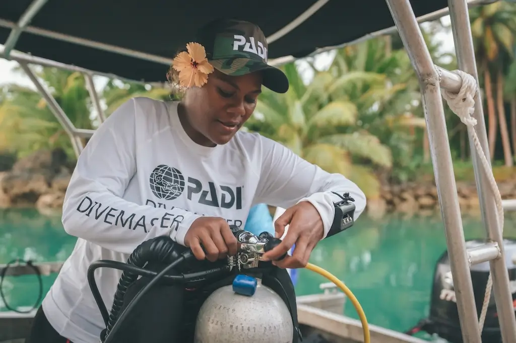 A divemaster on a boat checks scuba diving equipment and an air tank, wearing a PADI shirt and cap with a flower tucked behind her ear