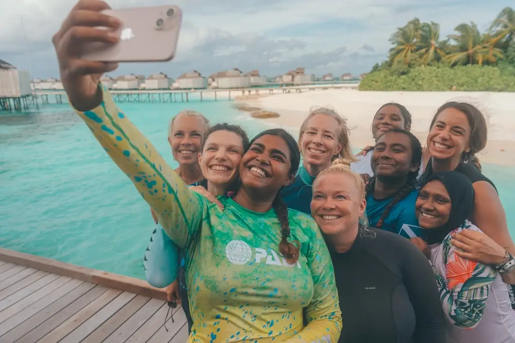 Group of scuba instructor students smiling together while taking a selfie on a tropical beach, with turquoise water, palm trees, and overwater villas in the background