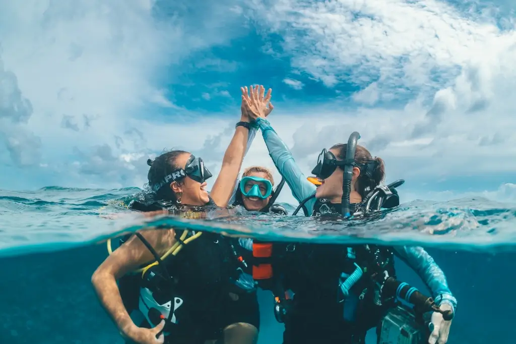 Two IDC students and their instructor celebrate a successful course dive with a high-five at the ocean’s surface, with their dive gear visible above and below the waterline.