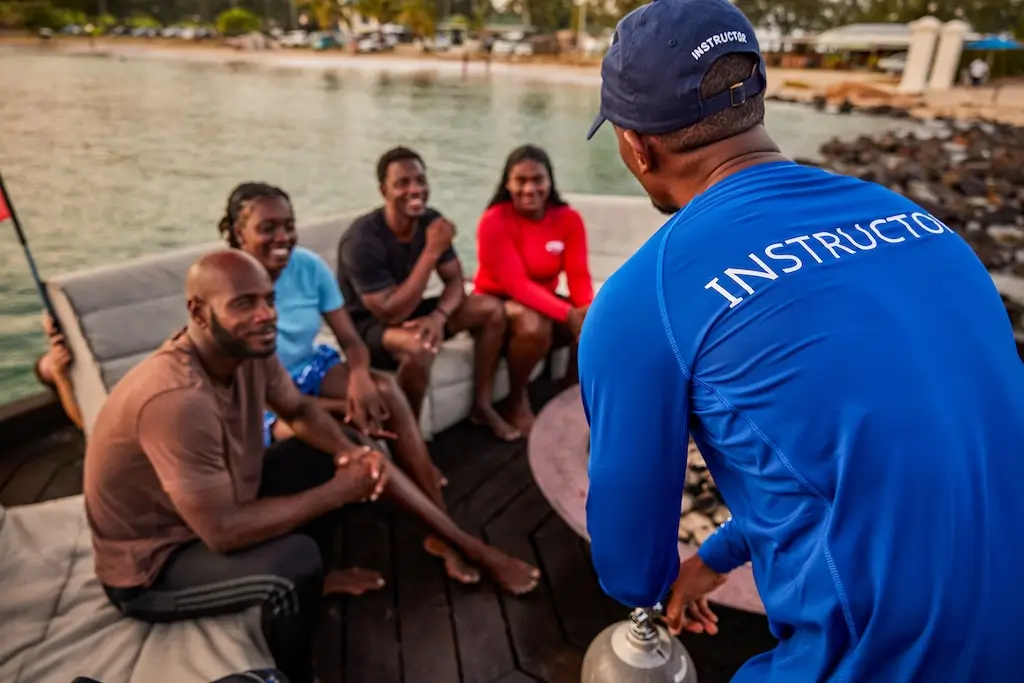 IDC students sit together on a boat dock beside the water while their instructor gives a briefing, with scuba equipment and a dive tank ready for training.