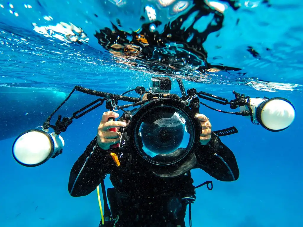 Underwater photographer in a black wetsuit holding a large camera housing with two lights, just below the water’s surface