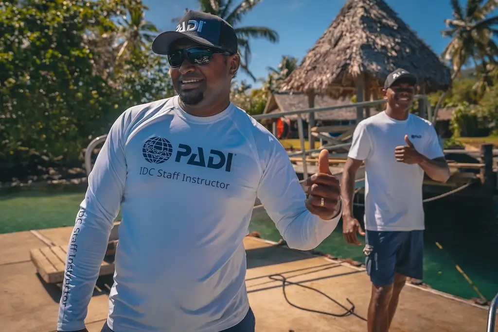 PADI IDC Staff Instructor smiling and giving a thumbs-up on a tropical dive boat dock, with another instructor and a thatched-roof structure in the background