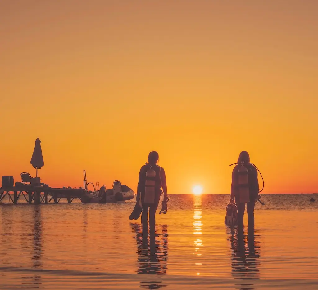 Two scuba divers stand waist-deep in calm ocean water at sunset, silhouetted against a vivid orange sky, with a dock and boats visible in the background