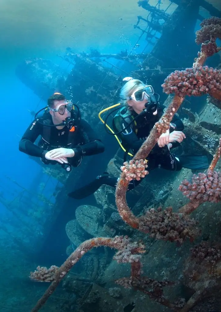 Two scuba divers explore a sunken shipwreck underwater, surrounded by coral-covered metal structures and beams in clear blue water